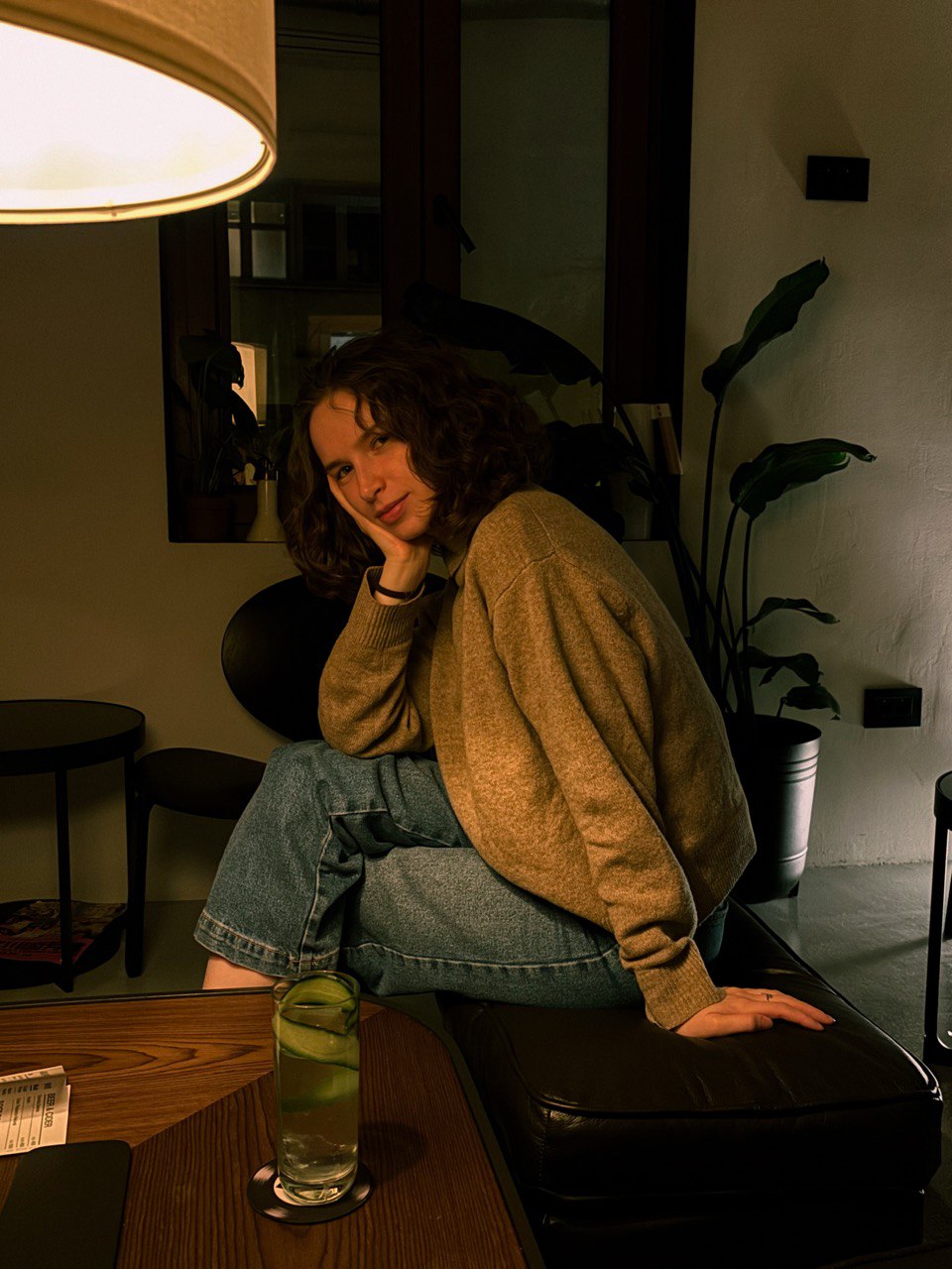 Katya in a beige sweater sitting on a dark bench, hand on chin.